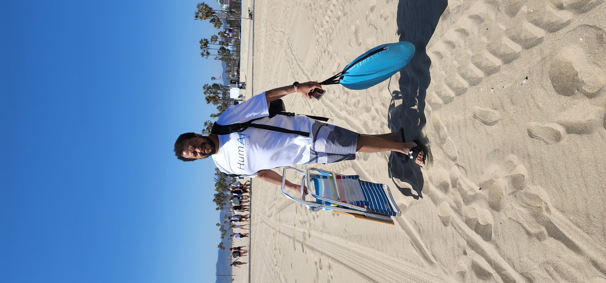 Rich walking across the sand at the beach carrying a folding chair and a beach bag